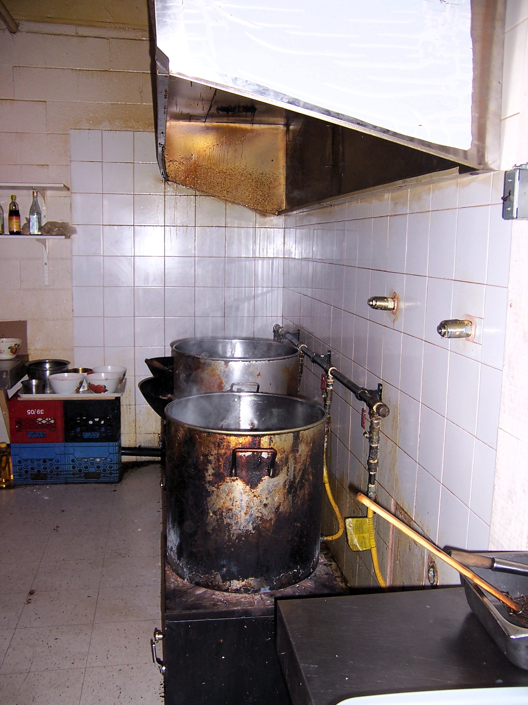 This image shows a commercial kitchen with large cooking pots under an exhaust hood. However, there are several fire and safety hazards present that should be addressed. Identified Hazards & Concerns:
⚠ Grease Buildup in Exhaust Hood – The accumulation of grease can pose a significant fire risk. Regular cleaning and maintenance are essential.
⚠ Burnt & Worn Cooking Equipment – The exterior of the cooking pots appears heavily charred, which may indicate overheating or poor ventilation.
⚠ Gas Line Safety – The exposed yellow flexible gas hoses should be properly secured and checked for leaks to prevent fire hazards.
⚠ Lack of Fire Suppression System – Commercial kitchens should have an automatic fire suppression system installed within the hood to control potential grease fires.
⚠ Poor Ventilation & Heat Control – Steam buildup suggests inadequate ventilation, which can cause overheating and increase fire risks. Recommended Actions:
✅ Deep Clean the Exhaust Hood – Remove grease buildup regularly to prevent fire hazards.
✅ Install & Inspect Fire Suppression System – Ensure the kitchen is equipped with a fire suppression system (e.g., wet chemical system).
✅ Check & Secure Gas Lines – Inspect gas connections for leaks and ensure all lines are properly fastened.
✅ Improve Ventilation – Verify that the exhaust fan system is functioning effectively to remove smoke, steam, and heat.
✅ Keep a Fire Extinguisher Nearby – Have a Class K fire extinguisher accessible for grease fires