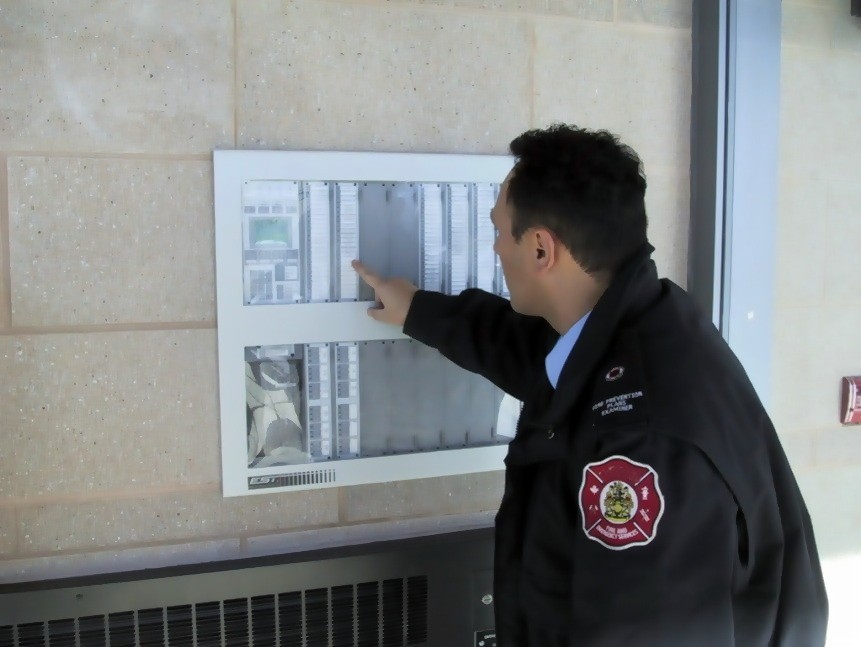 The image shows a fire prevention officer wearing a fire department uniform, inspecting an annunciator panel. The panel is mounted on a beige brick or stone wall and contains various labeled indicators and controls. The officer is pointing at a specific section of the panel, likely checking system status or troubleshooting an alarm. The setting appears to be a public or commercial building, given the professional fire safety inspection. Below the panel, a ventilation grille is visible, and to the right, a small red fire alarm pull station is mounted on the wall. The image suggests an official fire safety inspection or system check to ensure proper functionality of the building's fire alarm system.