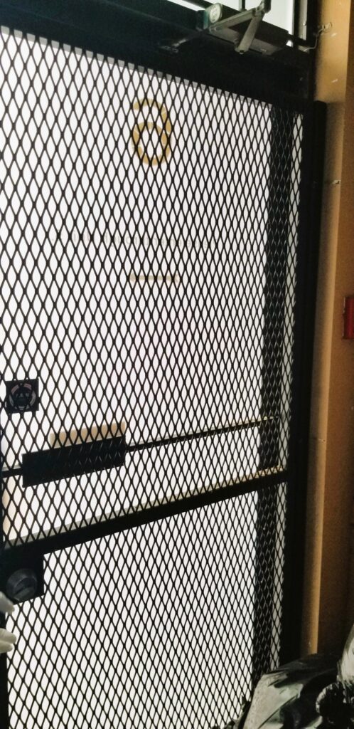 The image depicts a metal mesh security gate obstructing an exit door. The presence of a barricade like this can significantly hinder egress during emergencies, violating fire safety regulations. Emergency exits must remain unobstructed and easily accessible to ensure a safe evacuation route. This image likely highlights a fire code compliance issue.