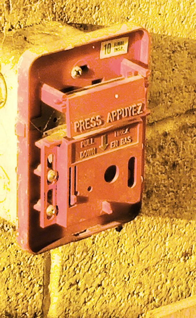 The image shows a damaged fire alarm pull station mounted on a wall. The device appears to be missing key components, which could prevent it from functioning properly in an emergency. Fire alarm pull stations are critical for initiating an alarm and alerting occupants of a fire hazard. A broken or inoperative pull station poses a significant safety risk and should be repaired or replaced to ensure compliance with fire safety regulations.