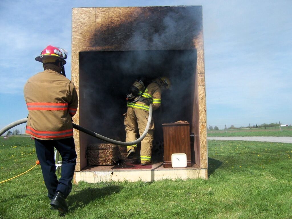The image shows two firefighters, dressed in full firefighting gear including helmets and protective clothing, engaged in what appears to be a training exercise or a controlled burn. One firefighter is standing outside a small structure, holding a hose, while the other firefighter is inside the structure, which is filled with smoke. The structure is made of wood and has a charred appearance at the top, indicating exposure to fire. Inside the structure, there is a small wooden cabinet and a white object that looks like a clock or a timer. The scene is set outdoors on a grassy area with a clear sky in the background. This image highlights the training and preparedness activities of firefighters, showcasing the importance of their role in fire safety and emergency response.