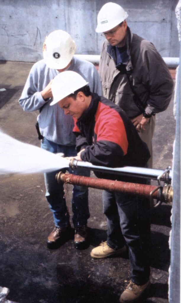 The image shows three men wearing white hard hats conducting a water flow test at an industrial or construction site. One man in the foreground, wearing a red and black jacket, is taking a pitot tube reading of pressure while the water is flowing forcefully by the fire pump. Water is forcefully flowing out of the hose, indicating a pressure test or system inspection. The other two men, one in a gray sweatshirt and jeans and the other in a dark jacket, are observing the process closely and recording readings. The setting appears to be an outdoor industrial area with concrete surfaces and some pipes visible in the background. The scene suggests a professional inspection or maintenance activity related to fire protection systems.