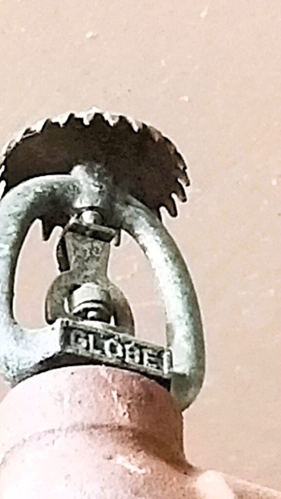 The photo shows a close-up view of an upright fire sprinkler head manufactured by "GLOBE." The sprinkler head is metallic and appears aged, with signs of corrosion. The deflector plate, which helps distribute water in a spray pattern, has a serrated circular shape. The sprinkler is mounted on a pipe, likely part of a fire suppression system in a building. The background is a beige or light-colored ceiling. This type of fire sprinkler is an essential component of automatic fire protection systems, designed to activate when high temperatures trigger the internal mechanism, releasing water to suppress fires.