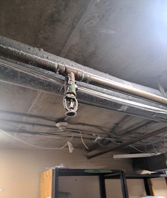This image shows a fire safety hazard related to a compromised sprinkler system. Key Fire Hazards Identified 🚨
🔴 Corroded & Dirty Sprinkler Head – This may prevent proper water discharge in case of a fire.
🔴 Obstruction by Dust & Debris – Buildup can delay activation or reduce water flow, leading to ineffective fire suppression.
🔴 Possible Pipe Damage or Leaks – The surrounding pipes show signs of wear, which could indicate potential failure. Recommended Actions:
✔️ Clean or Replace the Sprinkler Head – Ensure it is free of debris and fully operational.
✔️ Inspect the Fire Sprinkler System – A qualified fire protection technician should check for blockages, corrosion, or leaks.
✔️ Schedule Regular Maintenance – Implement a routine cleaning and testing program to ensure all sprinklers are in working condition.
✔️ Verify Compliance with Fire Codes – Ensure the sprinkler system meets NFPA 25 (Inspection, Testing, and Maintenance of Water-Based Fire Protection Systems)