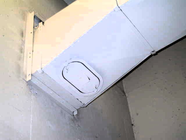 The image shows a close-up view of a white rectangular duct, likely part of an HVAC (Heating, Ventilation, and Air Conditioning) system. The duct is mounted on a wall and features a rectangular access panel with rounded corners and a handle for maintenance or inspection. The metal duct is securely attached to the wall with brackets, and the overall setting suggests an industrial or building environment focused on air circulation or ventilation systems with a fire damper mounted in the duct to prevent the spread of fire and smoke. The access panel provides access to the fire damper to be inspected.