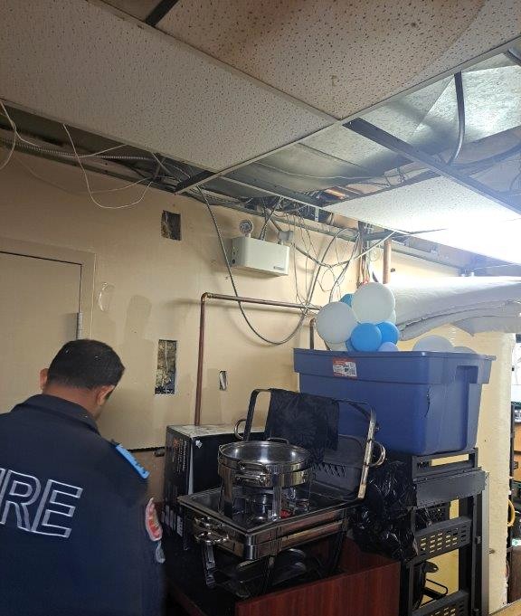 This image appears to depict a fire safety inspection in progress, and several potential fire hazards are visible.
Identified Fire Safety Concerns:
1. Exposed Wiring & Ceiling Panels
The open ceiling with hanging wires poses a fire risk.
Loose or exposed wiring could spark and ignite nearby materials.
2. Blocked Electrical Panel or Fire Equipment
The cluttered storage setup might obstruct fire suppression systems or electrical panels.
In an emergency, this could slow response time.
3. Improper Storage of Flammable Items
The presence of balloons (potential ignition hazard) and plastic storage bins near electrical equipment increases the risk of fire spread.
4. Cooking Equipment in an Unsuitable Location
The chafing dish and food warmer near electrical wiring and flammable materials may violate fire safety codes.
5. Wall Fire Separation
Wall is not continuous to the underside of the floor above
Recommended Actions:
Secure and properly enclose exposed wiring to prevent sparks.
Remove clutter near fire safety equipment to ensure accessibility.
Store flammable materials safely, away from ignition sources.
Ensure compliance with local fire codes regarding cooking equipment placement.
A follow-up inspection may be required to confirm compliance and mitigate fire risks.