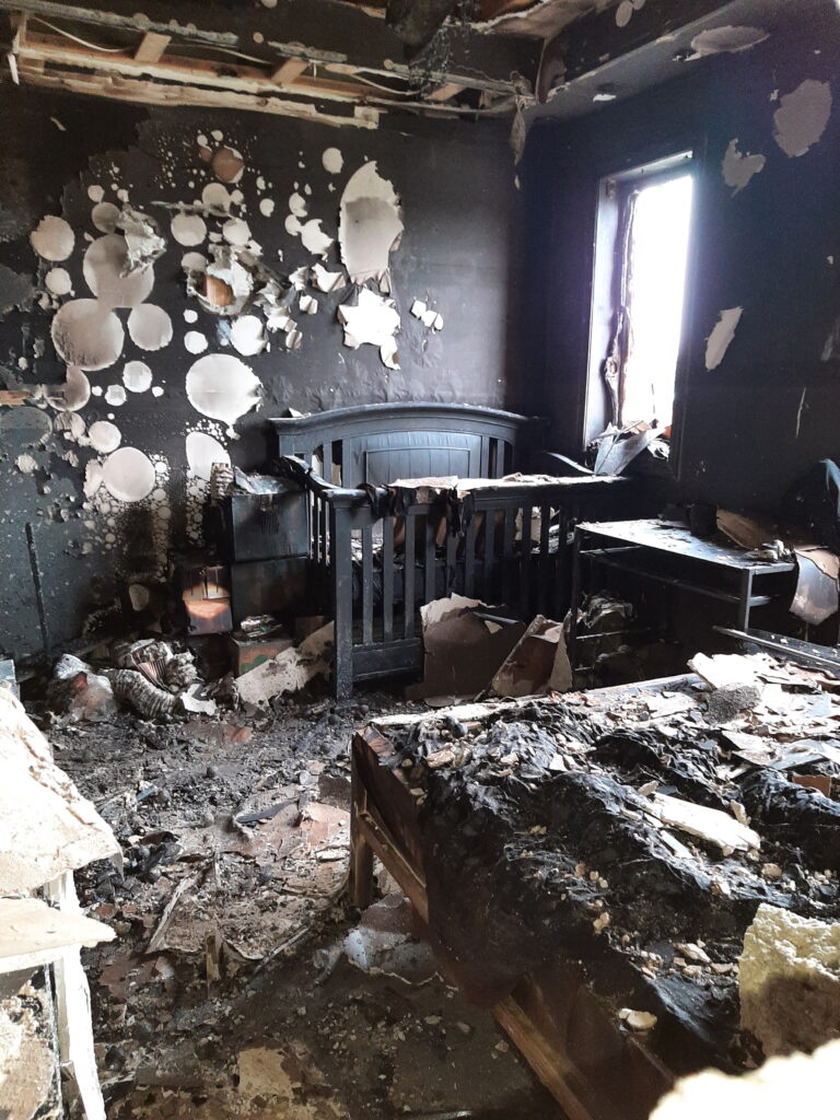 The photo shows a severely fire-damaged bedroom with extensive destruction. The walls are heavily blackened with soot, and large portions of the drywall have bubbled and peeled away due to extreme heat exposure. The ceiling is partially collapsed, revealing burnt wooden beams and structural damage. In the center of the room, a crib with a charred frame is visible, indicating that the space may have been a nursery. Nearby, a bed is heavily burned, with its mattress and bedding reduced to ash and debris. The floor is covered in fire-damaged materials, including remnants of furniture, insulation, and ceiling fragments. A window in the background appears to have been shattered or melted due to intense heat, allowing daylight to illuminate the destruction inside. The room's overall condition suggests a severe fire, likely requiring a thorough investigation into the cause and extent of the damage.