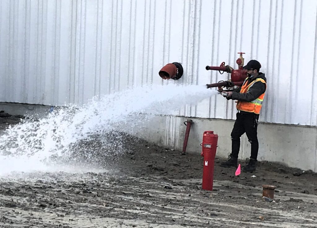 The image shows a worker in a high-visibility safety vest and protective gear conducting a fire pump test. He is operating the fire test header, releasing a powerful stream of water from a red play pipe mounted on the exterior of a large industrial or commercial building. The surrounding area appears to be a construction site or an area with unfinished ground. This test ensures that the sprinkler & standpipe systems are functioning correctly and providing adequate water pressure and sufficient water quantity for fire emergencies.