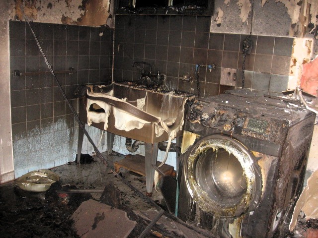 This image shows a fire-damaged laundry or utility room with severe charring and heat damage. The washing machine and sink are extensively burned, with melted plastic components and black soot covering most surfaces. The walls and ceiling exhibit signs of intense heat exposure, with peeling paint and structural damage.
A thorough fire investigation would be necessary to determine the root cause. Additionally, fire safety measures, such as proper ventilation, maintenance, dryer exhaust duct cleaning, circuit protection, and fire suppression systems, should be reviewed to prevent future incidents.