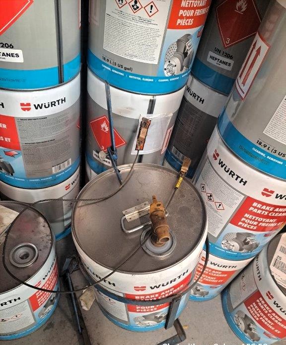 The image shows multiple metal barrels labeled "Würth" and containing brake and parts cleaner, specifically heptane, a highly flammable liquid. The barrels have hazardous material symbols, including a flame icon indicating flammability. One barrel in the foreground has a pump attachment with connected cables and clips, possibly for grounding or dispensing purposes. The setup appears to be in an industrial or workshop setting, with multiple stacked barrels stored together. This configuration suggests a potential fire hazard if not handled properly.