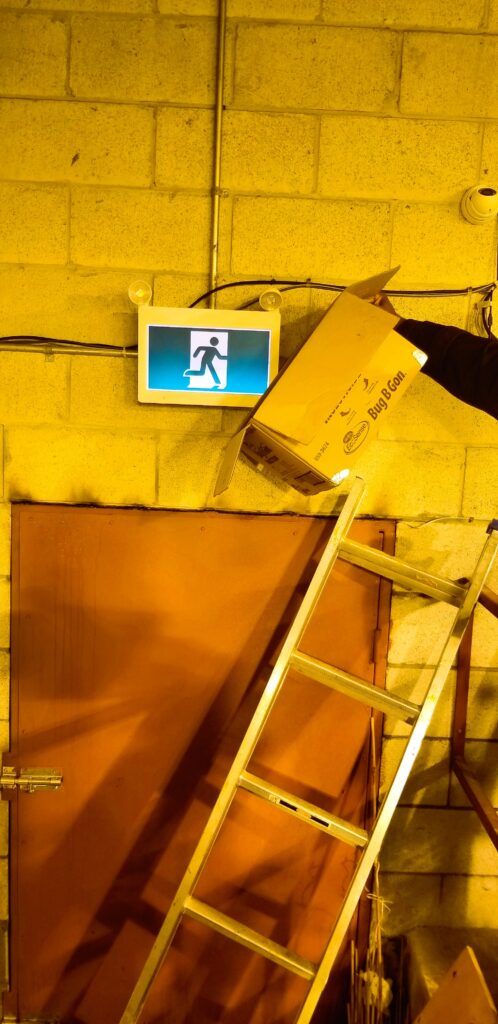 The image shows an illuminated emergency exit sign mounted on a concrete block wall. The sign features a running figure indicating the direction of an emergency exit. However, a cardboard box labeled "Bug B Gon" is being held in front of the sign, partially obstructing its visibility. A metal ladder leans against the wall near a brown door with a latch, which appears to be an exit obstructed by the ladder. The lighting in the area is yellowish, possibly from industrial or warehouse-style lighting. The image suggests a safety hazard due to the obstruction of the emergency sign, which could hinder visibility in an actual emergency.