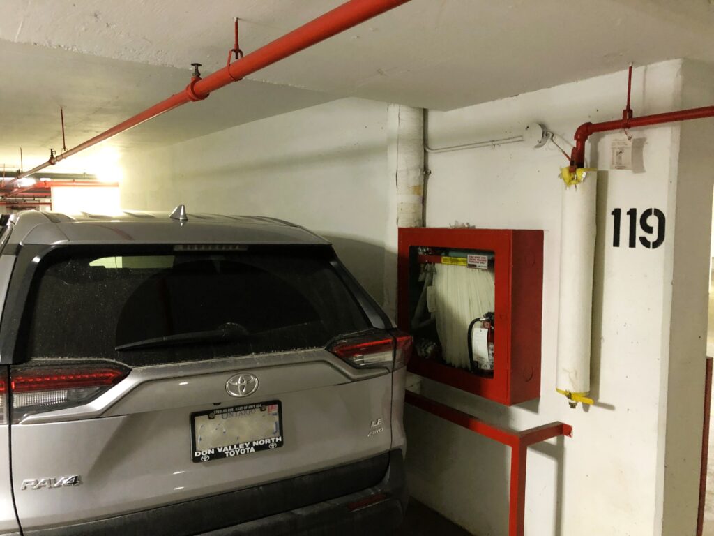 The image shows a silver Toyota RAV4 parked in an underground parking garage. The vehicle is positioned close to a red fire hose cabinet, partially obstructing access to it. The parking spot is marked with the number "119" on the white wall. Red fire sprinkler pipes run along the ceiling, and there is a white pipe with yellow tape attached to the wall. The garage is lit with artificial lighting. The car has a dealership frame from "Don Valley North Toyota," indicating it is registered in Ontario.