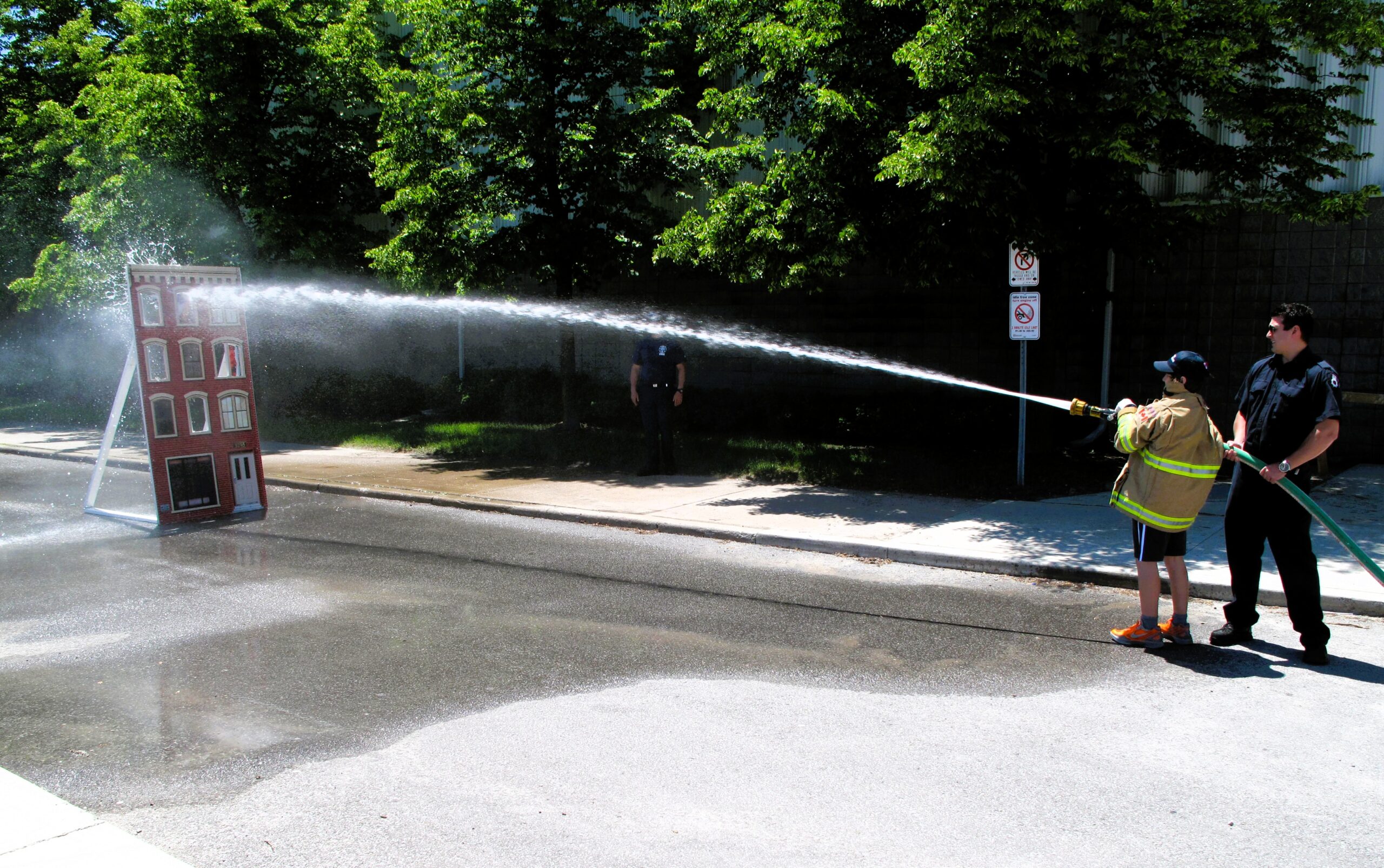 This image captures a firefighting demonstration at what appears to be a community or fire department open house event. A firefighter is assisting a young participant in using a fire hose to spray water at a mock building target. The event is likely an educational activity designed to engage the public, particularly children, in fire safety awareness and firefighting techniques. The bright outdoor setting and presence of safety personnel emphasize a hands-on learning experience in a controlled environment.