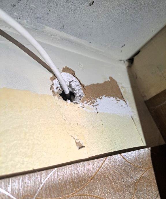 The image shows a white cable passing through a rough, unsealed hole in a wall or ceiling corner. The hole appears to have been manually drilled or chiseled, with exposed drywall, chipped paint, and uneven edges. The surrounding area has multiple layers of paint or plaster, some of which are peeling. The background includes a section of patterned wallpaper and a partially visible ceiling, which has dust or dirt accumulation. The installation appears unfinished or improvised, is a fire hazard because the unsealed hole will allow smoke and fire spread to other areas of the building. The hole requires to be fire stopped (sealed) to maintain the integrity of the fire separation.