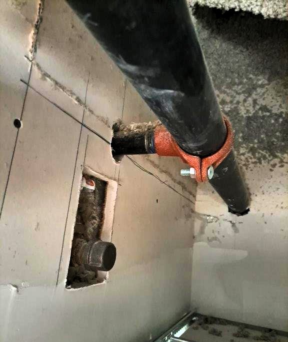 The image shows a close-up of a wall penetration where black sprinkler pipes pass through a drywall fire separation partition. The openings around the pipes are not properly sealed, and firestopping material appears to be missing or inadequate. Proper firestopping is crucial to maintaining the fire resistance rating of the wall and preventing the spread of smoke and flames in case of a fire. The image likely represents an inspection or assessment of firestopping compliance in a building.
