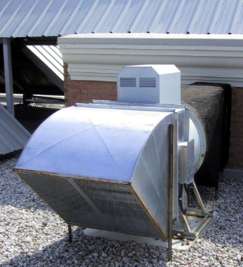 The photo shows a rooftop-mounted mechanical ventilation system, specifically a stairwell pressurization fan. The unit is housed in a metal enclosure with an angular intake or exhaust vent, designed to regulate airflow in a building's stairwell or shaft. The fan is secured on a metal frame and positioned on a gravel-covered rooftop. The surrounding structure includes a metal-clad roof with brick elements, indicating a commercial or multi-story residential building. The black protective cover on the duct suggests insulation or weatherproofing measures. This type of fan is commonly used for fire safety, ensuring that stairwells remain smoke-free in case of a fire by maintaining positive pressure, thereby allowing occupants to evacuate safely.