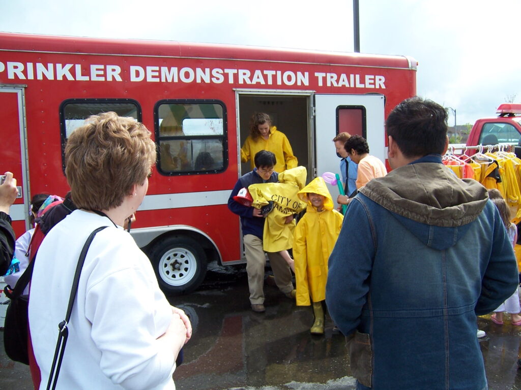 The photo shows a public fire safety event centered around a red Sprinkler Demonstration Trailer. Several people, including children and adults, are gathered around the trailer. Children wearing bright yellow raincoats and boots are exiting the trailer, likely after participating in a hands-on sprinkler system demonstration. Some of them are carrying yellow bags labeled "City of..." (possibly part of a fire safety program). Adults, including event organizers and spectators, are watching, taking photos, and engaging with the activity. A clothing rack with additional yellow raincoats is visible on the right side. The event appears to be an educational demonstration on how fire sprinklers work, likely aimed at raising fire safety awareness.
