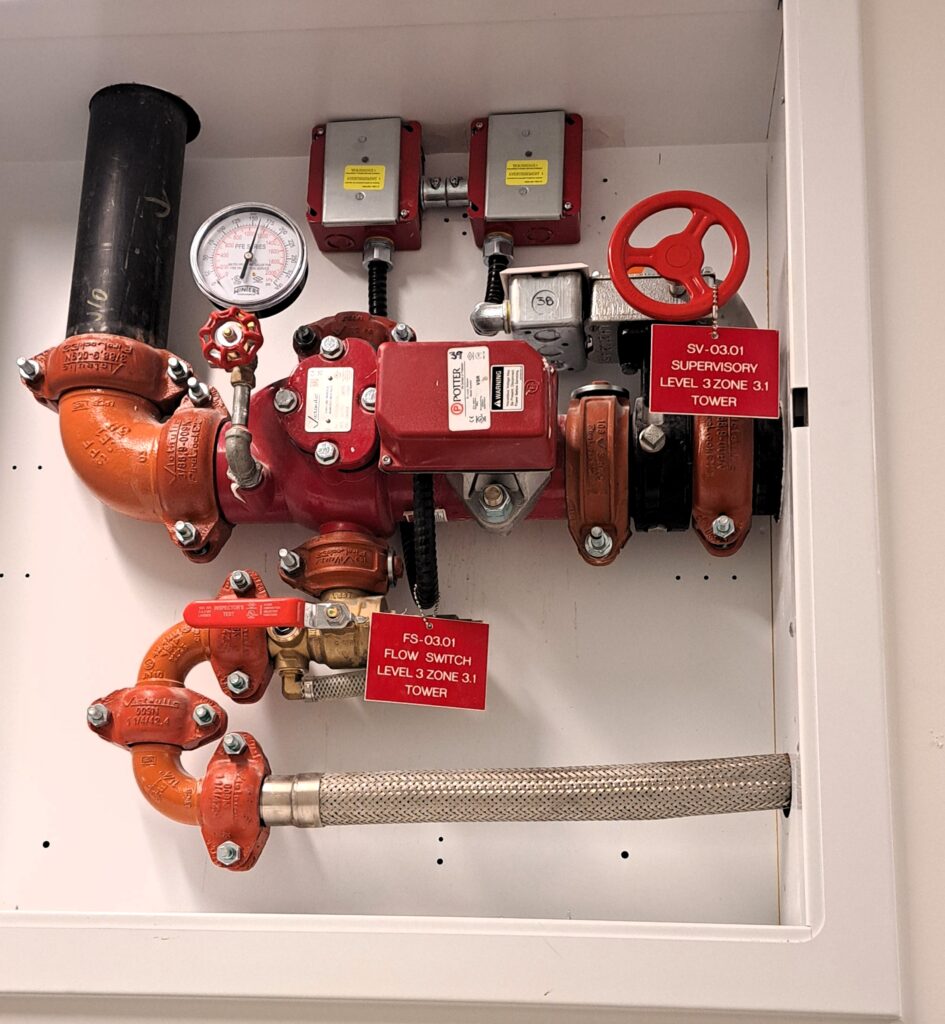 The photo shows a fire sprinkler system valve assembly inside a white enclosure. The system includes a red control valve (butterfly valve) with a circular handwheel, pressure gauge, flow switch, gauge and supervisory switches. The pipes are connected with orange grooved couplings, and the system has red identification tags indicating its function and location (Level 3, Zone 3.1, Tower). The flexible braided hose at the bottom is likely for drainage purposes. This setup specifically controls the 3rd level of the building is part of a building's fire protection sprinkler system, ensuring proper water flow and monitoring for fire suppression.