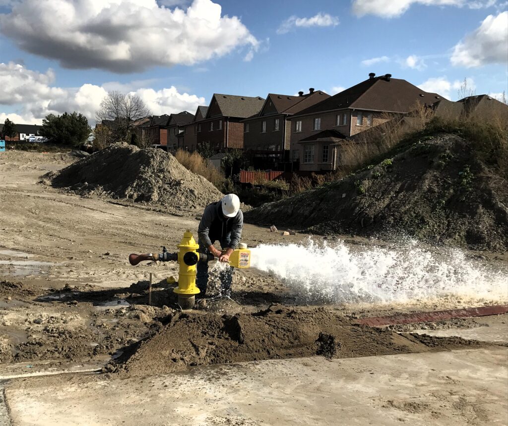 The image shows a construction site with a person wearing a white hard hat and a grey jacket operating a yellow fire hydrant. The person appears to be performing a water flow test, as a large stream of water is gushing out from it. The surrounding area is muddy and there are piles of dirt and construction materials visible. In the background, there are several residential houses with brown roofs and a clear blue sky with some clouds. The scene captures a moment of maintenance or testing of the fire hydrant, which is an essential part of urban infrastructure.